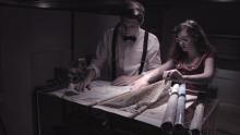 A screen grab of Lottie and Joe leaning over a tall table covered with several maps.