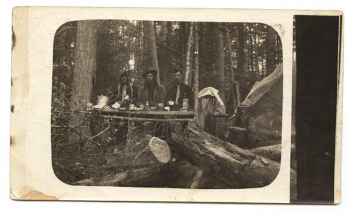 Une photographie noir et blanc de cinq hommes partageant un repas près d’une tente de prospecteur dans les bois.