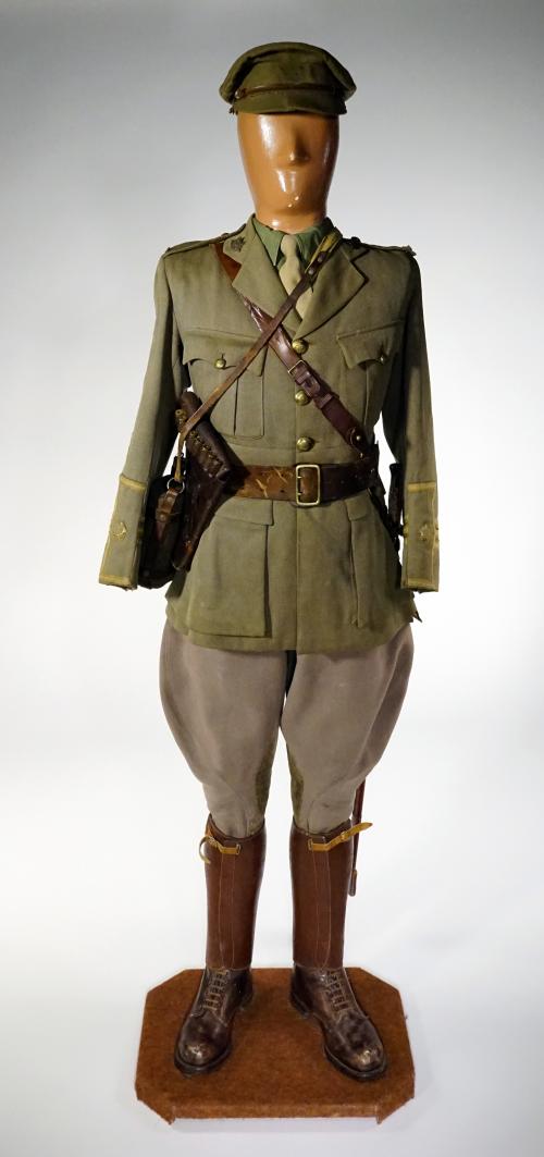 A mannequin is wearing a green khaki cap, a sword, a leather side arm holster and tunic; its cuffs are embellished with three stripes.