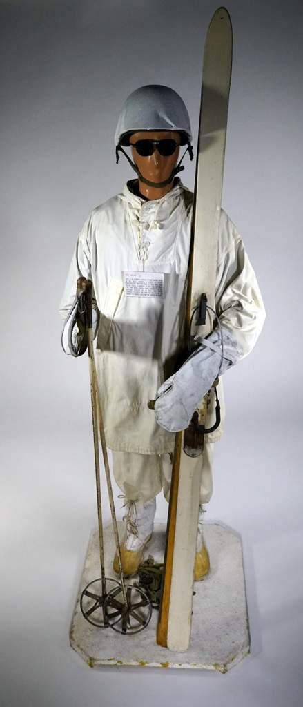 Un mannequin vêtu d’un uniforme militaire de camouflage d’hiver  portant des lunettes protectrices et tenant des skis dans ses mains.
