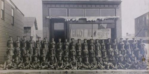 A black and white photograph of a large group of soldiers posing outside a  recruiting station.