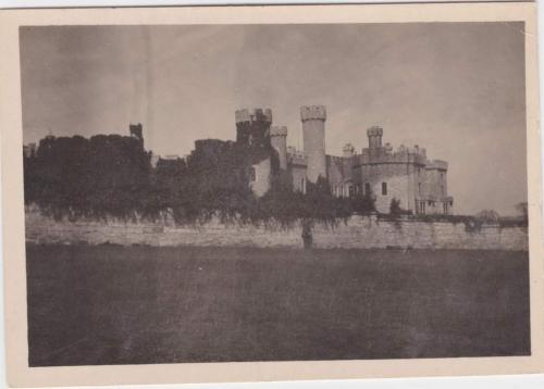 Une photographie d’un soldat posant devant un haut mur de pierres. On aperçoit un château en arrière-plan.