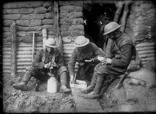Three Canadian soldiers are sitting in a trench opening their parcels and  enjoying a bite to eat.
