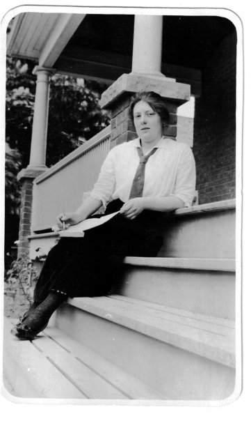 A black-and-white photograph of a young woman in a long skirt sitting on a set of stairs.