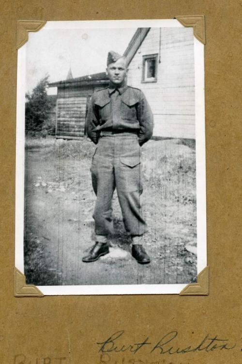 A black-and-white photograph of a soldier in uniform posing in front of a house.