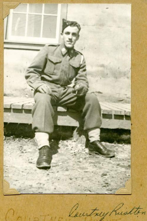 A black-and-white photograph of a young soldier in uniform sitting on a wooden sidewalk.