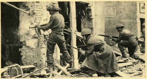 Une photographie en noir et blanc de soldats avec des pistolets fouillant des édifices bombardés.