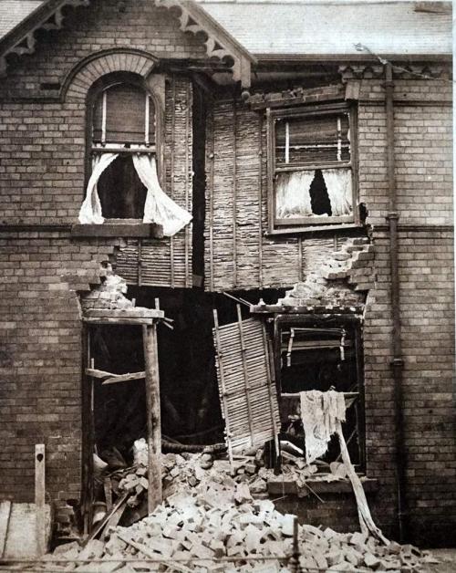 Une photographie d’un bâtiment de deux étages qui a été bombardé et dont la porte d’entrée a été détruite par une bombe.