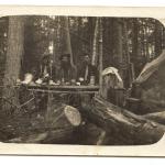 Une photographie noir et blanc de cinq hommes partageant un repas près d’une tente de prospecteur dans les bois.