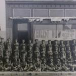 A black and white photograph of a large group of soldiers posing outside a  recruiting station.