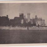 Une photographie d’un soldat posant devant un haut mur de pierres. On aperçoit un château en arrière-plan.