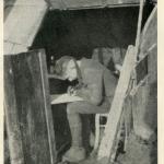 A black-and-white photograph of a soldier sitting inside a trench writing a letter.