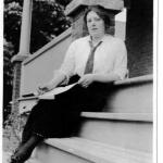 A black-and-white photograph of a young woman in a long skirt sitting on a set of stairs.