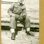 A black-and-white photograph of a young soldier in uniform sitting on a wooden sidewalk.