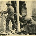 Une photographie en noir et blanc de soldats avec des pistolets fouillant des édifices bombardés.