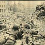 A black-and-white photograph of two soldiers lying with a PIAT at the end of a bombed-out street.